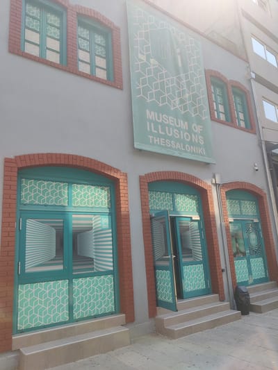 This media file shows the exterior of the Museum of Illusions in Thessaloniki, Greece. The building has a grey facade with red brick accents around the windows and doorways. The windows are tall and narrow, with teal frames and patterned glass. The lower panels of the doors are also decorated with a geometric teal and white pattern. The doors themselves are teal and some are open, revealing an optical illusion inside that creates a sense of distorted perspective. A large teal banner hangs on the building's facade with a geometric design and the text "MUSEUM OF ILLUSIONS THESSALONIKI" in white lettering. The sky is bright, suggesting it is daytime, and the lighting indicates sunny weather, with a slight glare visible in the upper right corner. The scene depicts the entrance to a museum. Visible text includes "MUSEUM OF ILLUSIONS THESSALONIKI". There are no people visible in the image.