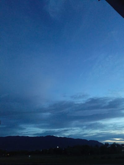 A horizontal shot captures the twilight sky over a dark silhouette of mountains and trees. The sky is a deep, clear blue, transitioning to lighter shades near the horizon where wispy clouds streak across. The lower third of the image shows the dark, undulating outline of mountains, with a line of trees in front of them. The foreground is a dark, indistinct area, possibly a field or road, with some faint lights visible in the distance. The scene is devoid of any people or discernable human activity. The overall impression is one of natural, serene beauty during the transition from day to night. The location is specified as Meunasahmanyet, Indonesia.