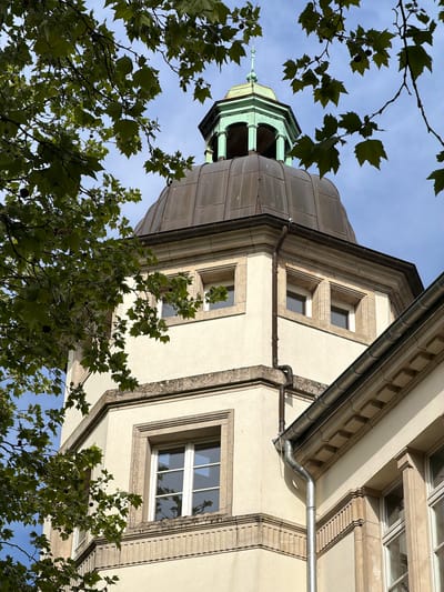 The image depicts an upward angle view of a building in Esch-sur-Alzette, Luxembourg. The structure features a tower with a copper-colored dome that transitions to a green cupola at the top. A spire-like element crowns the cupola. Parts of the building facade are visible, exhibiting a cream-colored stucco with light-colored stone framing windows. A metal gutter system runs along the edge of the building. The sky is visible in the background, with a blue hue interspersed with some cloudiness. Tree branches and leaves partially obscure the view, adding a natural foreground element to the architectural scene.