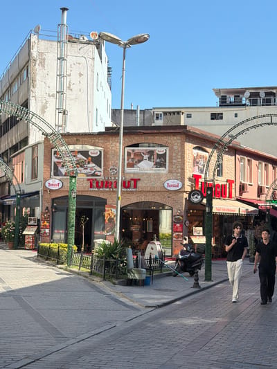 This media file shows a street scene in Istanbul, Turkey. The primary focus is on a restaurant called "Turgut Cafe & Restaurant," which features traditional Ottoman cuisine. The building has a rustic stone facade with large signs displaying the restaurant's name and advertisements for Turkish food, including images of traditional meals and French fries.
Two young men are visible in the foreground, one standing and the other sitting on a motorcycle. They appear to be casually dressed, with one wearing a black t-shirt and light-colored pants, and the other in a black t-shirt and black pants. Their presence suggests a relaxed atmosphere, possibly a break or a moment of rest.
The street is paved with brickwork, and a lamppost is prominent on the left side of the image. Decorative metal arches with lights are strung across the street, indicating a festive or commercial area. Greenery, including potted plants and hedges, is integrated into the restaurant's storefront and along the sidewalk, adding to the ambiance.
The time of day appears to be daytime, with bright sunlight casting shadows on the street, suggesting a clear and warm day. The blue sky visible in the upper portion of the image further supports this observation. The scene depicts a typical urban environment in Istanbul, blending historical architecture with modern commercial activity. Visible text includes "TURGUT," "RESTAURANT," "OSMANLI MUTFAĞI & KEI," and "Semah Gösterisi Free Dervish Show."