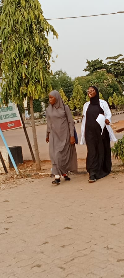 Two women are walking side by side on a dirt road with trees and a building in the background. The woman on the left wears a gray hijab and long robe, carrying a blue bag. The woman on the right wears a black dress, black hijab, and a white lab coat, holding papers in her hand. Both women appear to be of African descent. They are walking away from the camera. A sign with an arrow pointing left is visible behind the woman in gray.