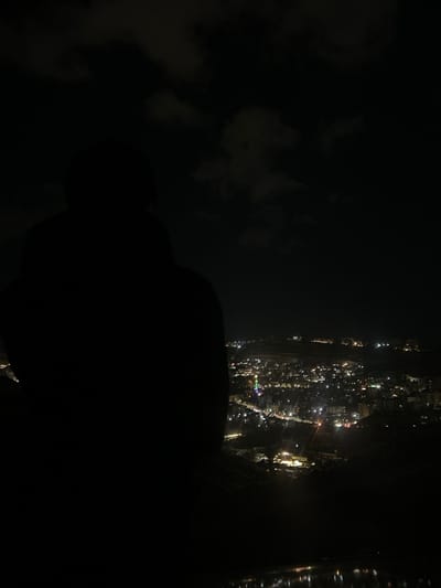At night, a cityscape glows with scattered lights seen from above. The view is partially obscured by dark, amorphous shapes in the foreground, likely plane elements, creating a sense of looking out of a window. Wisps of clouds dot the left side of the image, contrasting with the intense darkness. The city appears densely packed with illuminated buildings.