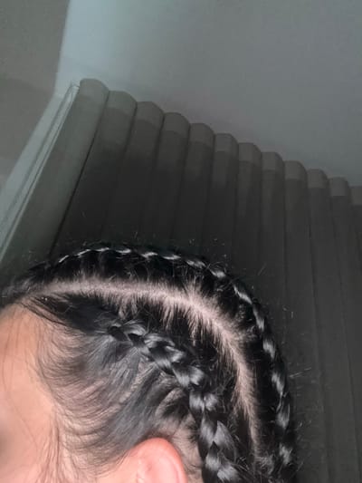 The image displays a close-up, angled view of the right side and top of a person's head, featuring dark hair styled into multiple cornrow braids. The braids run from the front hairline towards the back of the head, revealing visible sections of light-toned scalp between each braid. Fine, dark hairs are present along the temple and hairline. A portion of a flesh-toned ear is visible in the lower right quadrant. The background is out of focus, depicting a muted grey interior with vertical elements consistent with closed window blinds or curtains. The scene is illuminated by diffused light. This image is observed in the city of Kuklen, Bulgaria.