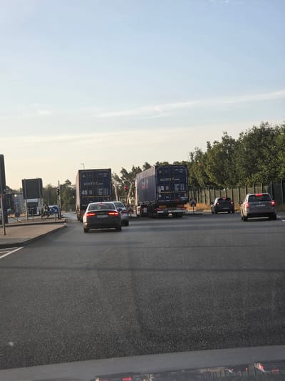 The image shows a traffic scene at a road junction, likely near a border crossing or port area, given the presence of container trucks. Two large blue shipping containers, branded "UNIT45.COM" and displaying the numbers "45" and "5", are prominently featured. One container is on a truck in the foreground to the left, and the other is on a truck further back. Several cars are visible, including a dark sedan with illuminated taillights in the left lane and a silver station wagon to its right, both appearing to be stopped or moving slowly. A smaller dark SUV is visible behind them.
In the background, trees line the horizon behind a green sound barrier fence. There are traffic lights, some illuminated red, indicating a stop. A person on a bicycle is seen near the traffic lights on the left side of the road. The sky is clear and pale blue, with a hint of yellow from the setting or rising sun, suggesting the photograph was taken during either dawn or dusk. The road surface is asphalt, with lane markings visible. The overall impression is one of a busy logistical area with active transport of goods.