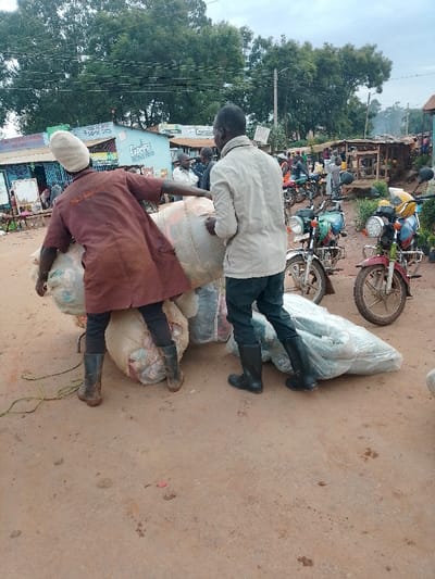 The image shows two men loading large, wrapped bundles onto a motorcycle. The men are standing on a dirt road in front of a small store, and the road is lined with other motorcycles and motorbikes. The man in the foreground, wearing a brown jacket, is lifting a large bundle onto the motorcycle. The man behind him, wearing a beige jacket, is helping to secure the bundle. The background is a dense urban area with multiple structures and vehicles, some in motion.  The scene appears to be a typical trading activity in the busy area of Kivaywa, Kenya, located at coordinates 0.562 latitude and 34.805 longitude. The scene suggests a bustling marketplace with commerce and trade occurring.