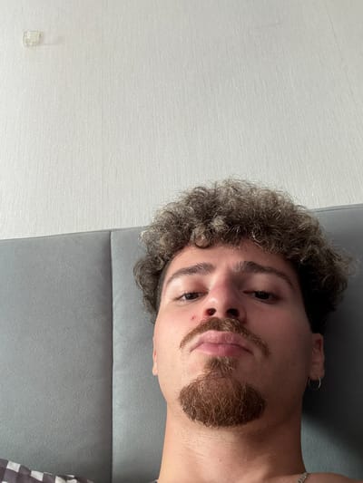 The image shows a close-up, low-angle selfie of a young man with curly, dark hair and a mustache and goatee. He is wearing a silver chain necklace and a hoop earring in his left ear. He has a red mark on his nose. The man is lying down, leaning against a grey headboard, with a textured, off-white wall behind him. There is a small, clear plastic object attached to the wall in the upper left corner of the frame. The scene appears to be indoors, likely in a bedroom. There is no indication of the location being in the city of Sao Thong Hin Town Municipality, Thailand.