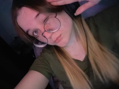 This image presents a close-up, angled portrait of a young female subject located in the city of Pushchino, Russia. The subject has long hair, dark brown at the roots and fading to a lighter blonde or ombré towards the ends, which falls over her shoulders and chest. She is wearing round, wire-rimmed eyeglasses and a dark olive green short-sleeved t-shirt. A thin silver-colored chain necklace is visible around her neck. A black headset is worn on her head, partially obscured by her hair and her raised left hand. Her fingers are positioned near her forehead, seemingly adjusting the headset or her hair. Her face is illuminated by a predominant reddish-pink light, highlighting her fair complexion. Her eyes are light-colored, and she maintains direct eye contact with the camera. Small dermal marks are present on her left cheek. The background is dark and out of focus, indicating an indoor environment with low ambient lighting.