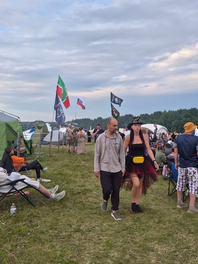 In Lagunovo, Russia, a festival setting is captured on a cloudy day. A group of people are seen in the foreground, with a man and a woman walking towards the camera. The woman is wearing a black top, a red and black tutu skirt, a black hat adorned with accessories, and a yellow fanny pack. The man beside her is in a light gray hooded shirt and dark pants. To their right, another man in a blue t-shirt and checkered shorts walks away from the camera. In the background, tents are pitched on a grassy field, and more people are gathered. Several flags are visible, including a green, red, and white flag, a black flag with a skull emblem, and a Russian flag. Some individuals are seated in camping chairs, and a water bottle is placed on the ground next to one of them. The overall scene suggests a relaxed outdoor gathering or event.