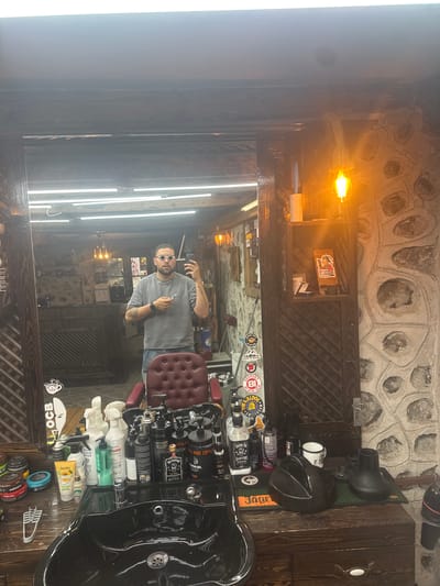 The image captures a reflection in a mirror inside what appears to be a barber shop. The man reflected in the mirror is taking a picture with a cell phone. The shop features a stone wall, wooden details, and rows of fluorescent lights. A barber's chair and products such as gels, sprays, and pomades are visible in the foreground. Stickers are displayed on a wooden shelf. The text suggests that the location may be Bansko, Bulgaria.