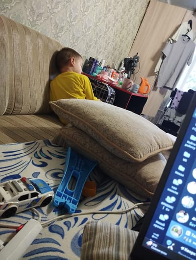 The image depicts an indoor scene, likely a living room or bedroom, with a young boy in a yellow shirt sitting on a couch. The couch has a striped pattern. In the foreground, there are stacked pillows with a patterned design. To the left, a toy police car is visible, along with a white power strip with a red indicator light. A blue plastic object and an orange object are also on the floor. The background shows a wall with patterned wallpaper, a shelf with various items including cups, and a gray t-shirt hanging on a hanger. A laptop screen is partially visible in the lower right corner, displaying what appears to be a messaging application with profile pictures and text. The scene does not provide any specific visual cues to pinpoint its location within Kingisepp, Russia.