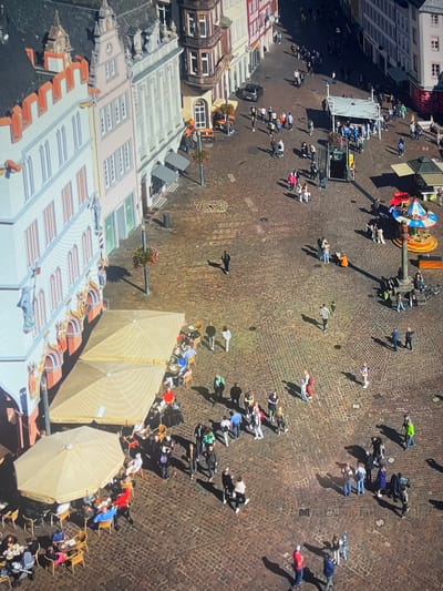 The image shows an overhead view of the Hauptmarkt in Konz, Germany, with a brick-paved market square bustling with activity. Buildings with distinct architectural styles surround the square, including a row of buildings with a multi-layered, tiered roof design. A portion of the square is filled with numerous individuals walking in various directions, creating a lively pedestrian environment. Some people are gathered under umbrellas, suggesting a possible outdoor seating area for cafes or restaurants. Additionally, a small amusement ride can be seen in one corner of the square, adding to the vibrant atmosphere.