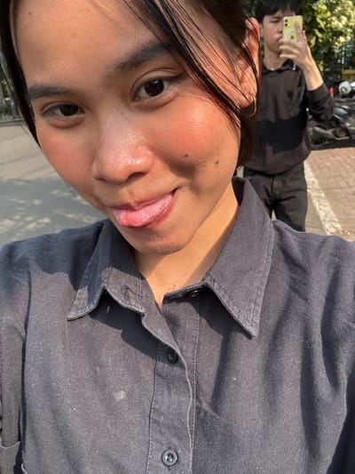 The media file shows a close-up, low-angle selfie of a person wearing a dark grey, collared shirt. The person's face is partially visible, with their tongue sticking out. Their skin appears shiny, suggesting it might be warm or humid. To the left of the main subject, another person is visible in the background, holding a yellow smartphone and seemingly taking a photo. This second person is wearing a black hoodie and dark pants.
In the background, a paved area with a white line is visible, along with some greenery and the side of a motorcycle. The sunlight suggests it is daytime, likely with clear weather. The shadows cast on the ground indicate a bright sun.
The setting appears to be outdoors in a place with some urban elements, given the paved ground and motorcycle. The overall impression is casual and relaxed. The activity appears to be taking a selfie, with another person also engaged in taking a photo. The time of day is likely midday due to the strong sunlight and shadows. The emotions conveyed are playful and lighthearted due to the tongue-out pose. No visible text is present in the image. The location context is South Jakarta, Indonesia.
