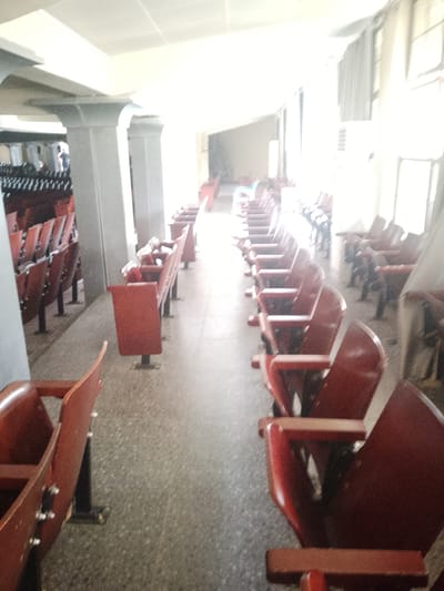 The image depicts an indoor seating area, likely a lecture hall or auditorium, with rows of empty, dark red seats. The seats are arranged in straight lines, receding into the distance. The flooring appears to be a speckled gray material. In the background, sunlight streams in from windows, creating a bright, hazy effect that obscures details. The overall impression is of an unoccupied public space. The specific location in the city of Kuson, Nigeria, cannot be determined from the visual information provided.