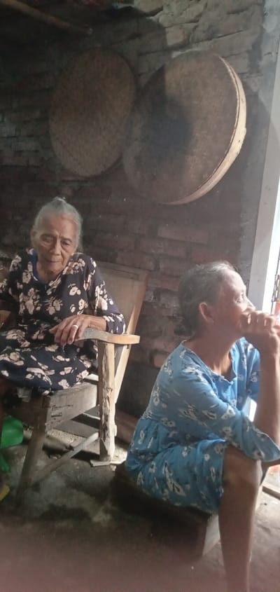 This image, captured in Baron, Indonesia, depicts two females inside a dimly lit room with an exposed red brick wall. An elderly woman, with gray hair pulled back, wearing a dark blue floral long-sleeved dress, sits on a worn wooden chair in the mid-left frame, looking directly forward. A silver-colored ring is visible on her left hand's ring finger. To her right, a second female, appearing middle-aged and also with dark hair pulled back, wearing a light blue floral long-sleeved dress, sits on a low wooden bench or platform. She faces towards the right side of the frame, looking out of a brightly lit opening, with her right hand resting near her chin or mouth. Two large, circular, woven basket-like objects hang on the brick wall in the background. The floor is an uneven, dark concrete or dirt surface.