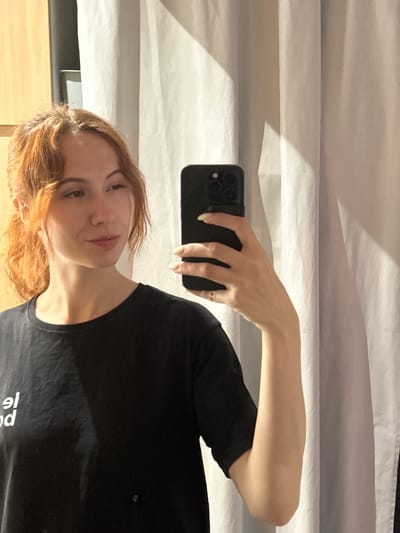 A young woman with auburn hair tied in a ponytail, wearing a black t-shirt with white text, takes a mirror selfie. She is holding a black smartphone in her right hand, which is visible in the frame. The background consists of a light-colored curtain and a wooden closet. The scene does not provide any specific information about its location in Novi Sad, Serbia.