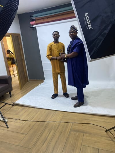 Two men stand on a white platform within a photo studio setting. The taller man wears a blue traditional robe and matching hat, while the shorter man is in a gold-colored suit. Both men face forward with serious expressions. Studio equipment, including a large softbox, are visible. A doorway in the background shows a glimpse of a room interior. The floor outside the platform is covered with light wood-tone planks.