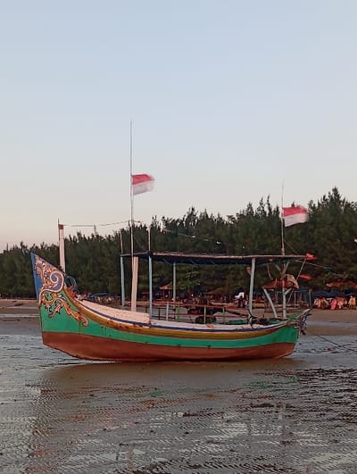 A traditional Indonesian fishing boat is beached on a wet, textured sandy shore in Tritunggal, Indonesia. The boat features a reddish-brown lower hull, a green upper hull with a yellow stripe, and an intricately carved and painted blue, gold, and green ornate prow. A simple canopy structure covers part of the boat. Two tall, thin poles extend from the vessel, each flying a small red and white Indonesian flag. The immediate ground surface shows ripple patterns and reflections. In the background, a dense line of dark green trees fronts a beach area where several open-sided huts and indistinct structures are visible. The sky is clear and pale blue. No individuals are clearly identifiable in the immediate vicinity of the boat.