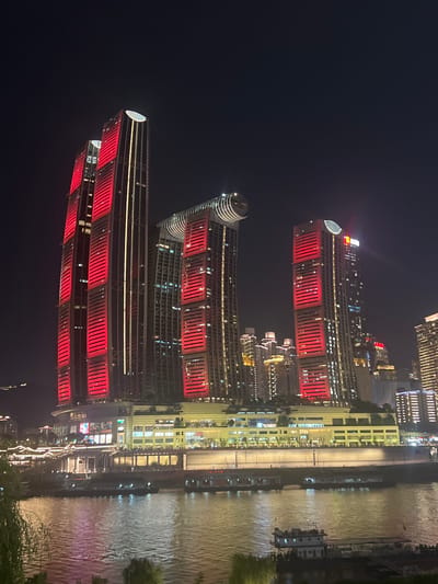 The image shows a nighttime cityscape with illuminated skyscrapers and a river in the foreground. The prominent structures are modern high-rise buildings, many of which are adorned with bright red and yellow lights, creating a vibrant display against the dark sky. One building, in particular, features a distinct curved element near its top. In the foreground, a body of water reflects the city lights. Several boats are visible on the river. The buildings appear to be part of a commercial or residential development, possibly along a waterfront promenade or urban waterfront area. The overall impression is one of a bustling, modern Chinese city at night. The location is identified as 江北城街道, China.