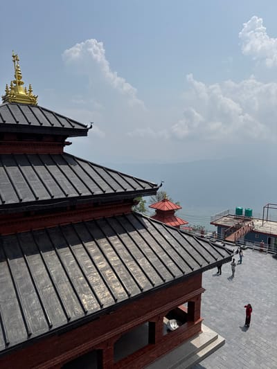 The image depicts a scene in Pokhara, Nepal, focusing on a multi-tiered building with a dark, sloped roof and golden accents. Above, a section of the sky is visible with scattered clouds. In the upper right corner, figures of people are discernible on a paved surface adjacent to the building, along with a structure with exposed pipes and tanks. The perspective is from a somewhat elevated position, tilted, emphasizing the height of the building and the aerial view of the city.