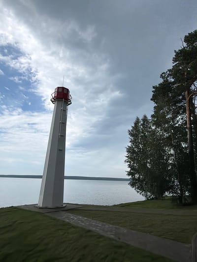 This is a medium-shot, eye-level, outdoor photograph taken on a cloudy day. A tall, white, conical lighthouse with a red top stands on the left side of the image. The lighthouse is positioned on a grassy area near the edge of a large body of water, which appears to be a lake or a wide river. A paved path leads towards the lighthouse from the lower foreground. To the right of the lighthouse, several trees, including tall pines, are visible along the shoreline. The water stretches to the horizon, where a distant treeline is visible. The sky is filled with dramatic, heavy grey clouds, with some patches of lighter sky and scattered white clouds showing through on the left. The scene appears to be a scenic overlook or a park area. The image is believed to have been taken in Zarechny, Russia, near the Belaia River. There are no people visible in the frame.