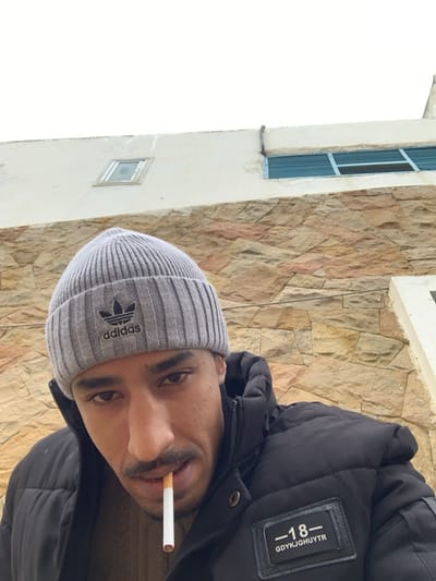 A male individual is depicted in a close-up, upward-angled shot against the exterior wall of a building in M'diq, Morocco. The subject is wearing a grey ribbed beanie with a black Adidas logo, a black puffer jacket with a rectangular patch displaying "-18-" and "GDYKJGHUYTR" in white text, and a light brown knitted garment underneath. A white cigarette with an orange filter is positioned between his lips. He has a mustache and is looking directly at the viewer. The background consists of a building wall, featuring a lower section with irregular light brown and grey stone cladding, and an upper section of a lighter, possibly off-white, painted surface. A small, square, recessed window is visible in the upper left background, and a horizontal window with blue horizontal slats is present in the upper right background. The sky, visible at the top, is overcast and bright.