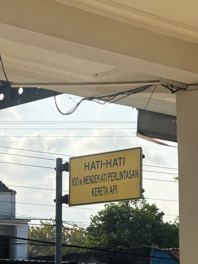 The image captures a close-up view of a yellow traffic sign in Kedawung, Indonesia. The sign, mounted on a metal pole, displays text in Indonesian: "HATI-HATI 100 M. MENDEKATI PERLINTASAN KERETA API". This translates to "CAUTION 100 M. APPROACHING RAILWAY CROSSING". The sign is positioned under an overhang of a building, with wires and conduits running above it. In the background, power lines crisscross the sky, which is partly cloudy. Trees and rooftops of buildings are visible at the bottom of the frame, suggesting an urban or suburban setting. The overall scene is devoid of people or any ongoing activity.