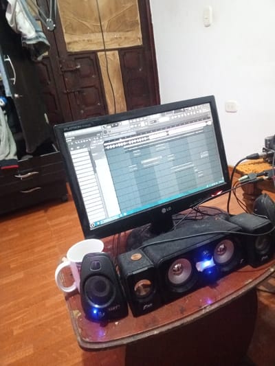 An LG monitor displays music production software on a wooden desk. Speakers, with glowing blue lights, are arranged in front of the monitor, alongside a white mug. The desk appears worn. In the background, there's a dark-colored wardrobe, drawers, and a white wall with an electrical outlet. A brown, paneled door is also visible. The room appears to be in a residential setting.