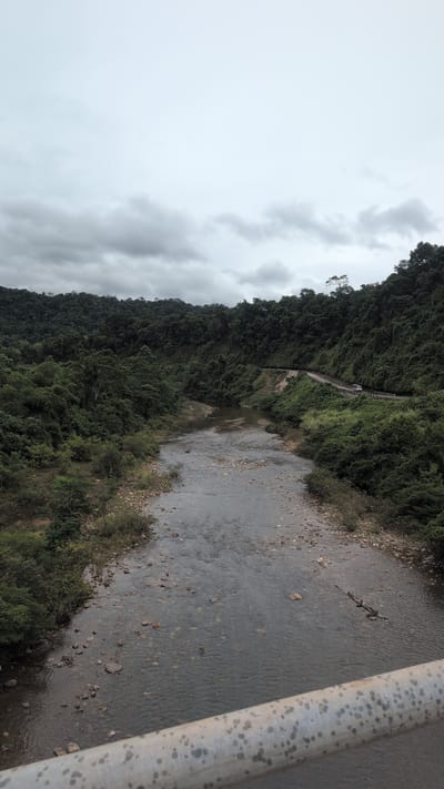 A wide, shallow river flows through a lush, densely forested valley under an overcast sky. The riverbed is visible with numerous stones and pebbles. On the right bank, a paved road winds along the hillside, with a white vehicle traveling on it. The forested slopes rise steeply on both sides of the valley, characterized by a dense canopy of green trees. The foreground shows the edge of a metal railing, possibly from a bridge. The scene is one of natural beauty and a rural landscape. The location is identified as Trường Sơn Commune, Vietnam.