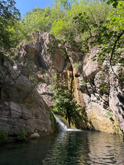 The image depicts a natural scene with a small waterfall cascading into a tranquil pool of water. The pool is surrounded by steep, rocky cliffs covered in lush green vegetation and trees. Sunlight filters through the leaves, creating dappled patterns on the rocks and water. The scene is serene and appears to be a remote natural location. The location is identified as Krapina, Montenegro.