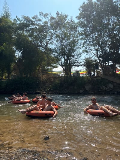 A group of people are floating down a river in orange inflatable tubes. Most are young men with bare chests. The water is clear but shallow, and there is greenery along the bank, including trees and shrubs. A building or structure is visible in the background, and a cloudy sky is above.