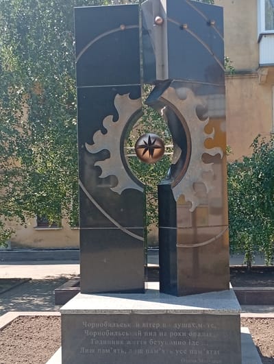 This is a medium shot, eye-level, outdoor photograph of a monument. The monument is made of dark granite and features intricate metalwork. In the center of the monument, there is a spherical object with a starburst pattern. The monument is surrounded by trees and foliage. In the background, a building with light-colored walls and windows is visible. The base of the monument has engraved text in Ukrainian. This monument is located in the city of Pokrov, Ukraine, and it is dedicated to the Chernobyl disaster. The text on the monument reads: "Chernobyl wind in souls, dust settles on us for years, life goes on endlessly... Only memory, only memory. Olena Matyshchuk".