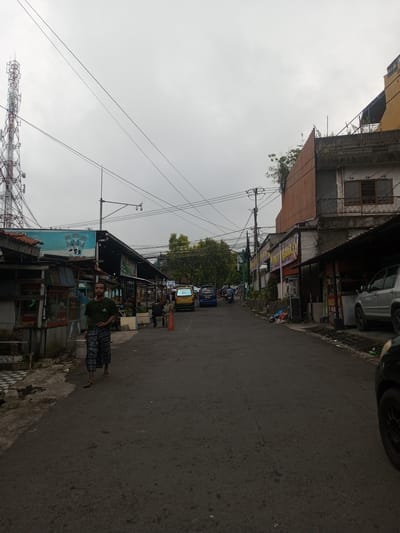 An overcast street scene in Houaythat, Laos, depicts a commercial and residential thoroughfare. In the immediate foreground, an asphalt road occupies the lower portion of the frame. On the left, a male subject with short hair and a beard walks towards the viewer, slightly off-center. He wears a dark green t-shirt with a white logo and blue-and-white plaid sarong-style pants, along with sandals. To his left, a damaged sidewalk with black and white checkerboard tiles and exposed earth is visible.
Along the left side of the street, a series of weathered, informal market stalls or small shops constructed from corrugated metal, wood, and concrete are present. Above these structures, a large blue billboard advertising "Kopi Good Day" is prominent. Further left and in the background, a tall red and white cellular/radio transmission tower rises against the grey sky.
On the right side of the street, the front wheel and lower body of a dark vehicle are partially visible in the bottom right corner. Adjacent to this, a silver SUV is parked next to a covered commercial structure displaying a "Bakso" sign. The buildings on the right range from single-story shops to multi-story residential or mixed-use structures, some with visible balconies and overgrown vegetation.
The midground of the street shows various vehicles, including a yellow van, a blue sedan, and several motorcycles, indicating active traffic. An orange traffic cone is positioned on the right side of the road. Multiple utility poles line the street, supporting a dense network of overhead electrical and communication wires. A street light assembly with a mounted camera is also visible. Several indistinct figures are present further down the street, walking or operating vehicles. Trees with green foliage are visible further down the street, providing some natural elements amidst the urban setting. The sky is uniformly grey and overcast.