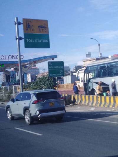 A daytime shot features a gray SUV driving on a road. Above the car is a sign indicating a "Toll Station" with "MTC" and "ETC" icons. Further ahead, a sign points to "Mombasa Rd" and "Expressway." A large bus is visible to the right, with people standing nearby. Barriers with yellow and black stripes line the roadside, and buildings are seen in the background under a clear sky.