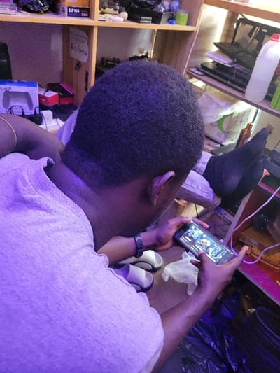 An individual, an adult male with short, dark hair, is depicted from an elevated rear-right perspective in an indoor setting, assumed to be in Makurdi, Nigeria. He wears a light-colored t-shirt, a gold-colored bracelet on his left wrist, and a dark watch on his right wrist. The scene is illuminated by a prominent purple ambient light source. The individual is actively engaged with a black smartphone, held horizontally with both hands, displaying what appears to be a mobile game or application interface. A pair of white and grey/black slip-on sandals and a crumpled white cloth are situated on a surface to his left. In the upper right background, a foot wearing a dark sock is visible, propped up. A light-colored wooden shelving unit occupies the background, containing various items including a box labeled "2.5" HDD CASE," PlayStation 5 controller packaging, several laptops, a white plastic bottle with a red cap, green cylindrical containers, and a brown glass bottle. The immediate floor area features dark plastic materials and visible power cables, including white and pink strands. The environment suggests a cluttered personal space, potentially a room dedicated to electronics or leisure.