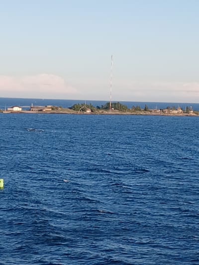 The image shows a body of water, likely the Baltic Sea, with an island in the distance. The island appears to have some buildings and vegetation, as well as a tall, striped antenna or radio mast. The water is blue and choppy, with small whitecaps. There are a few small white boats visible on the horizon. A bright yellow buoy is partially visible in the lower left corner of the image. The sky is clear and pale blue. The scene is likely located in the archipelago of Helsinki, Finland, a region known for its numerous islands and extensive coastline.