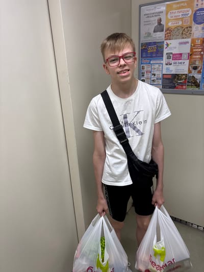 The image shows a young male wearing a white t-shirt with the Calvin Klein logo and black shorts, holding two plastic grocery bags filled with items. He is also wearing a black fanny pack across his chest and red-rimmed glasses. The background features a bulletin board with various advertisements, including pizza promotions and services. The bulletin board has text in Russian. One of the advertisements mentions "БИРМА" and "ПИЦЦЫ", which are Russian words for "stock exchange" and "pizzas" respectively. Another advertisement mentions "ЛЕТНИЙ ЛАГЕРЬ", meaning "summer camp". The overall setting appears to be a public space within the city of Kemerovo, Russia, possibly a shopping center or a public transit waiting area.