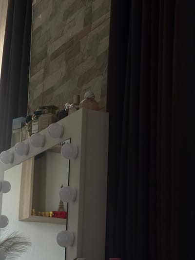 In Zmeitsa, Bulgaria, a dressing table with a mirror illuminated by light bulbs is positioned against a wall with textured stone tiles. On the top edge of the dressing table, several perfume bottles are arranged. The scene is framed by dark, heavy curtains on either side, suggesting an indoor setting, possibly a bedroom or dressing room. The mirror reflects a shelf with decorative items, including a miniature Eiffel Tower.