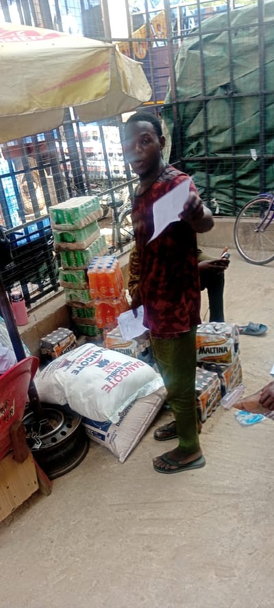 The image captures a bustling scene in what appears to be a market or trading area, likely in Fagge A, Nigeria, based on contextual clues. A man of African descent, clad in a dark t-shirt and olive-green trousers, stands in the foreground, holding a piece of paper and possibly speaking to the viewer. Merchandise is scattered around him including stacks of soft drinks, cases of Maltina (a malt drink), and bags of Dangote-branded goods (likely sugar and/or salt). The setting looks like a permanent trading spot, evidenced by the overhead awning and fenced-in area in the background. A bicycle is also visible. The overall environment suggests a busy marketplace.