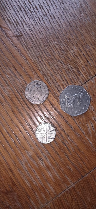 This image shows three British coins on a wooden surface. The coins are a 2009 fifty pence piece, a one pound coin with a shield design, and another coin that is partially obscured. The wooden surface appears to be a floor or tabletop with a prominent wood grain pattern. The lighting is from above, casting slight shadows on the coins. The location of Marum, Netherlands, is not depicted in this image.