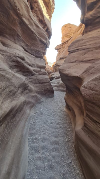 The image shows a view within the Red Canyon, located near Eilat, Israel. The perspective is from within the canyon looking upwards, showcasing the undulating, carved sandstone walls. The walls are layered with varying shades of brown and beige. The canyon floor is covered with small, gray gravel. A small opening at the top of the canyon wall reveals bright sunlight.