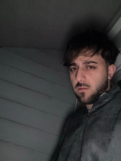A man with dark hair and a beard rests his head on gray paneling, possibly a wall or floor. He wears a gray jacket with a visible zipper. The lighting is dim, creating shadows that accentuate the texture of the paneling and the contours of his face. His expression is neutral, with his eyes looking towards the camera. The overall mood is subdued and intimate.
