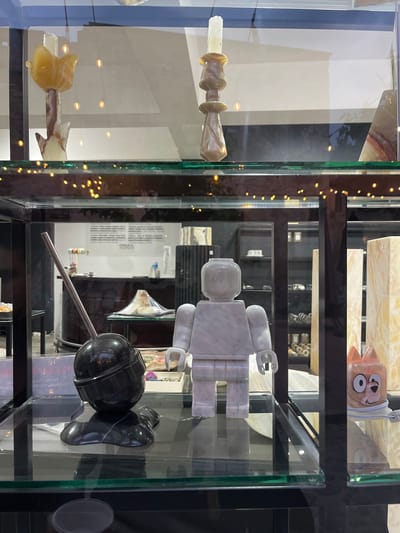 The image shows a display case in what appears to be a shop or gallery in Domingo Arenas, Mexico. The case is filled with various objects, likely artworks or decorative items. On the top shelf, there is a pink object with cartoonish features. The middle shelf holds a large white marble sculpture resembling a Lego figure. The bottom shelf contains a black sphere attached to a metal rod, possibly a sculpture, accompanied by a black object resembling a shoe. The overall lighting is bright, and the objects are displayed in a clear, organized manner.