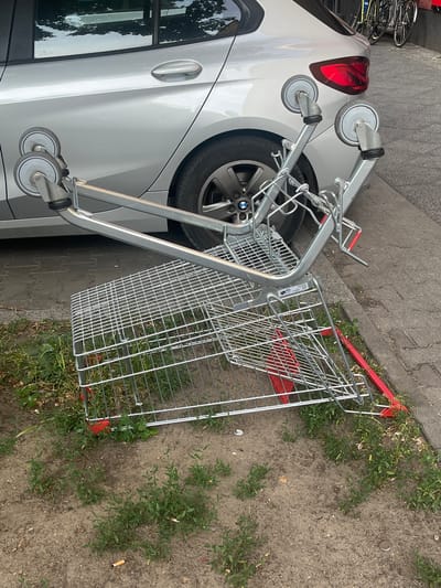 A silver BMW is parked on a sidewalk next to a shopping cart. The shopping cart, partially on the grass and sidewalk, appears to be either abandoned or in the process of being moved. The car is a hatchback, and its rear taillight is visible, which is red and angular. The car's wheel is also visible. The shopping cart is mostly metal with red plastic accents on its base and handles. It is tilted, suggesting it might have fallen over or is precariously balanced. The scene appears to be outdoors during the day, with sunlight casting shadows. The ground consists of pavement and patches of dry grass and dirt. In the background, more parked bicycles are visible, hinting at a urban environment. There is no discernible text in the image. The overall impression is one of a mundane urban setting, with the shopping cart adding a touch of disorder. The weather appears to be fair.
