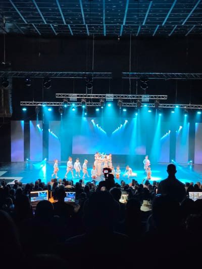 The image shows a stage performance with a group of young dancers on a brightly lit stage. The stage is decorated with vertical blue and purple panels that are illuminated by spotlights. The dancers, dressed in light blue and white costumes, are positioned in various poses, with some standing and others kneeling. In the foreground, an audience is seated in a darkened theater, with some individuals holding up phones to capture the performance. There is also a camera on a tripod positioned in the audience, aimed at the stage. Two laptops are visible in the lower part of the image, suggesting a technical setup for the event. The lighting creates a vibrant and dynamic atmosphere. The scene appears to be an indoor event, likely a dance recital or competition.