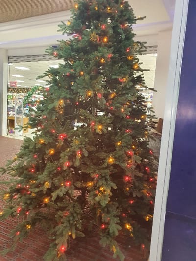 An artificial Christmas tree, approximately 2-3 meters in height, stands indoors within a commercial establishment. The tree is densely branched and illuminated by numerous small, evenly distributed red and orange/yellow lights. The floor beneath is covered with a patterned reddish-brown carpet or tile. In the background, a commercial storefront is visible, featuring a partially open grey, horizontally slatted roll-down security shutter. Beyond the shutter, the interior of a brightly lit retail space with shelving units and merchandise displays is faintly discernible. A light-colored drop ceiling with embedded lighting fixtures is visible in the background. A vertical white structural column is partially visible on the right side of the frame. No individuals are present in the scene. The specific location within Kisumu, Kenya, is not identifiable from the visual evidence provided.