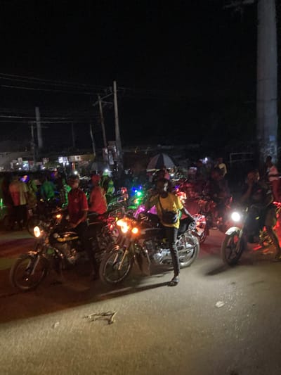 The image displays a crowded night street scene in Ura Nta Umuarandu, Nigeria. A large gathering of individuals, primarily male, are present, many positioned on or adjacent to numerous motorcycles. In the immediate foreground, a male subject wearing a yellow t-shirt, dark trousers, and dark sandals stands to the right of a dark motorcycle with its headlight illuminated; he holds a dark object, likely a mobile phone, to his right ear and wears a dark shoulder bag. To his left, another male subject in a red shirt and dark pants is seated on a similar dark motorcycle, also with an illuminated headlight.
Additional motorcycles, some with active headlights and others adorned with multi-colored LED string lights (blue, green, red), are visible throughout the frame. Further back, numerous people and motorcycles create a dense crowd. In the mid-ground, a person holds an open umbrella featuring distinct red and light-colored stripes. The background reveals blurred structures indicative of buildings or stalls lining the street, with multiple utility poles and overhead power lines silhouetted against the dark night sky. The ground surface is paved, possibly asphalt or packed earth, with some indeterminate debris in the closest foreground. Illumination is provided by vehicle headlights, decorative lights, and distant artificial sources, casting varied shadows across the scene.