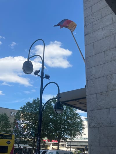 This is a vertically oriented, outdoor photograph taken from a low angle looking up at a bright blue sky with scattered white clouds. A street lamp with multiple lights and cameras is prominently featured in the foreground, its dark metal structure arching across the frame. A flag, with horizontal stripes of yellow, pink, and blue, is visible flying from a pole on the left side of the image.
In the background, a building with large windows and beige stone cladding is partially visible at the top. Further in the distance, lush green trees are in full leaf, suggesting a warm season, possibly summer. Below the trees, a street scene unfolds with glimpses of buildings, shops, and a yellow double-decker bus. Some text is visible on the buildings, including "ZO", "Gray Line", "LOGE", "PKN", "OBA", and "SHOPPEN SCHNAB' 'ERE". The overall impression is a sunny day in a city, likely Berlin, Germany, with a festive or pride-related element suggested by the flag. The low angle of the shot emphasizes the sky and the architectural elements above.