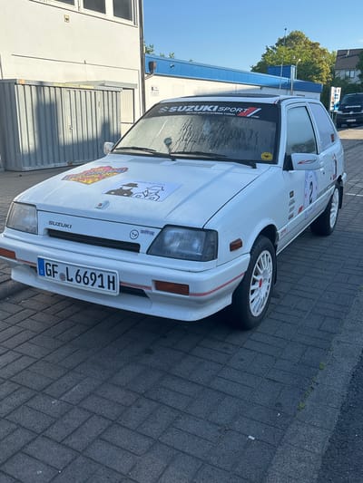 The image shows a white Suzuki Swift parked on a brick-paved surface. The car has racing decals on the hood and windshield, including "SUZUKI SPORT," "MIT HUT," and rally-themed stickers. The license plate reads "GF L6691 H," indicating that the car is registered in Germany, specifically the district of Fulda (GF). The windshield is slightly water-spotted. The wheels are white with red center caps. In the background, there's a building with silver siding and a partially visible dark-colored vehicle. The sky is clear and blue, suggesting daytime. The presence of a commercial-looking building suggests an urban or industrial area within Fulda.