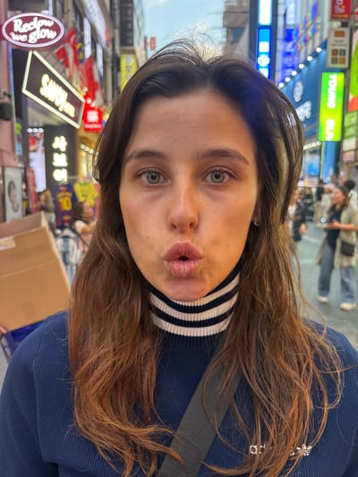 This is a selfie of a young woman with long, brown hair. She is wearing a blue sweater with a white and black striped collar. Her lips are pursed, and she is looking directly at the camera. She has light-colored eyes and fair skin.
The background appears to be a busy street scene, likely in Seoul, South Korea, given the visible text. There are many neon signs and lights, suggesting it might be nighttime or an area with abundant artificial illumination. The signs include Korean characters and some English words like "YOUNG OL" and "REDGLOW WE GLOW". The overall atmosphere seems vibrant and urban. The woman's expression is playful and engaging.