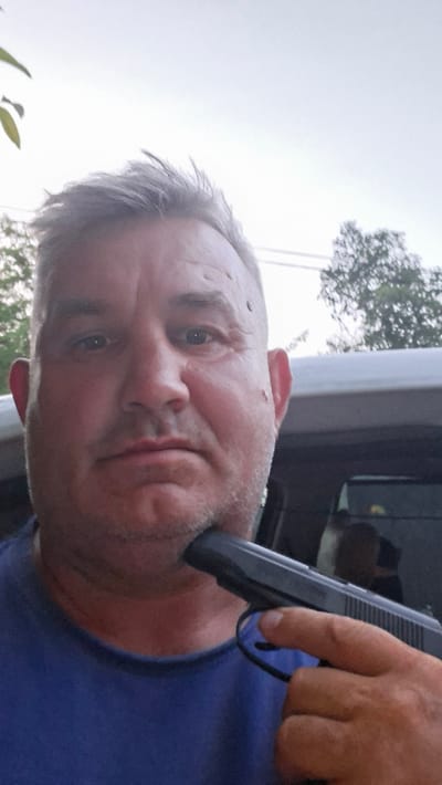 A man is seen holding a handgun to his chin in a selfie. He is wearing a blue t-shirt and has graying hair. The background is blurred but appears to be outdoors with some trees and a vehicle. The image is a close-up, focusing on the man's face and the weapon. There is no indication of the location being Voinova, Moldova.